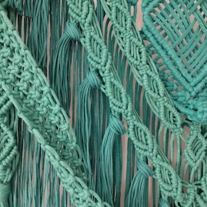 Op de afbeelding: Een tealblauwe macram&eacute; wandhanger met verschillende knoop patronen en texturen. De hanger heeft een groot vierkant patroon in het midden en franje details.