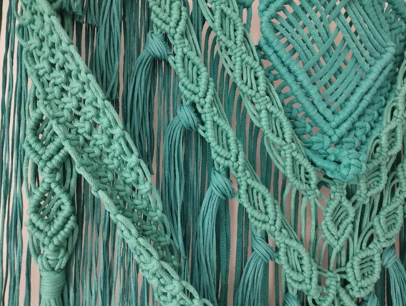 Op de afbeelding: Een tealblauwe macram&eacute; wandhanger met verschillende knoop patronen en texturen. De hanger heeft een groot vierkant patroon in het midden en franje details.
