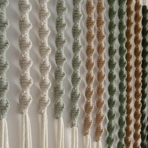 May include: A wall hanging made of macrame cord in various shades of beige, green, and brown. The cord is knotted into a series of loops, creating a textured and visually appealing design.