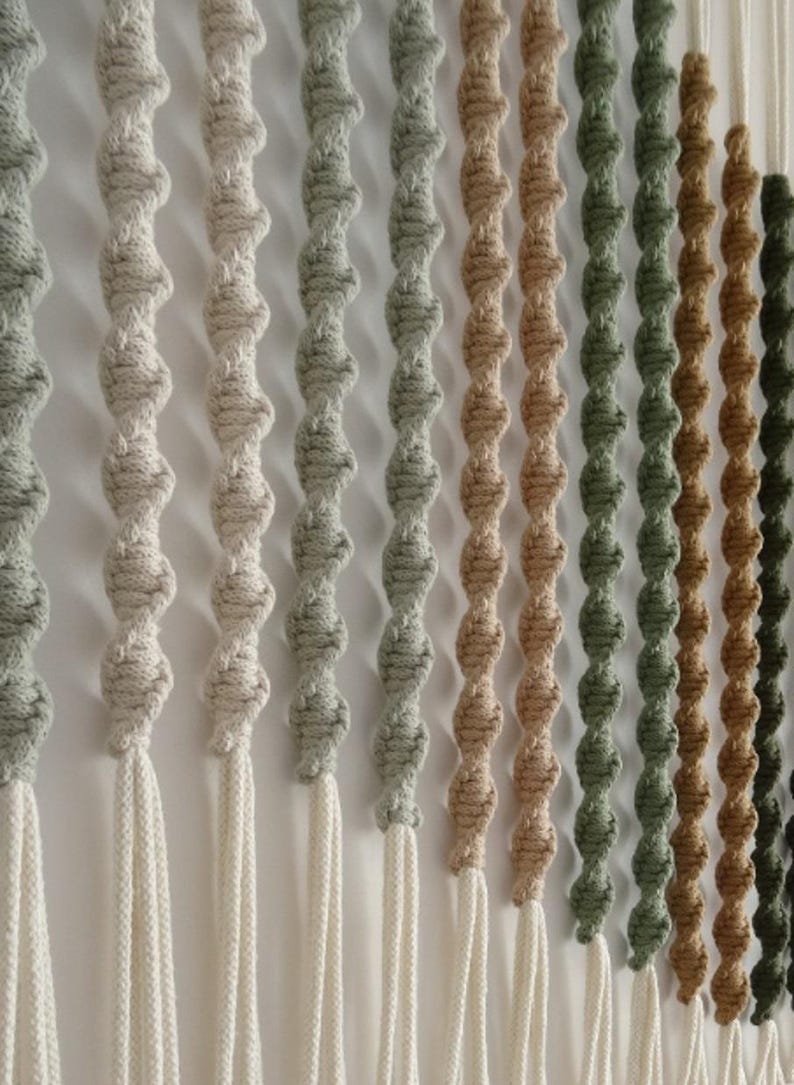 May include: A wall hanging made of macrame cord in various shades of beige, green, and brown. The cord is knotted into a series of loops, creating a textured and visually appealing design.
