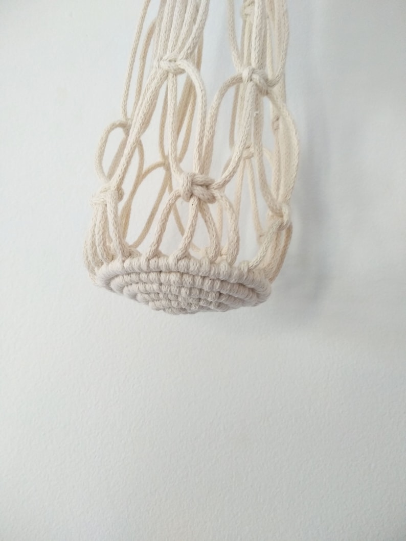 May include: A white macrame hanging planter with a circular base. The planter is made of thick cotton cord and has a woven design.