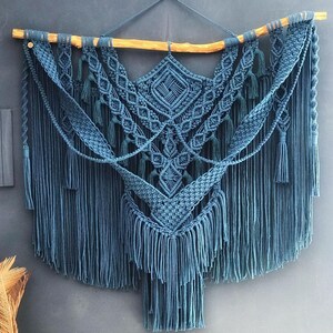 Op de afbeelding: Een grote, blauwe macram&eacute; wandhanger met een geometrisch patroon en franjes. De hanger is gemaakt van katoenen koord en is bevestigd aan een houten tak.