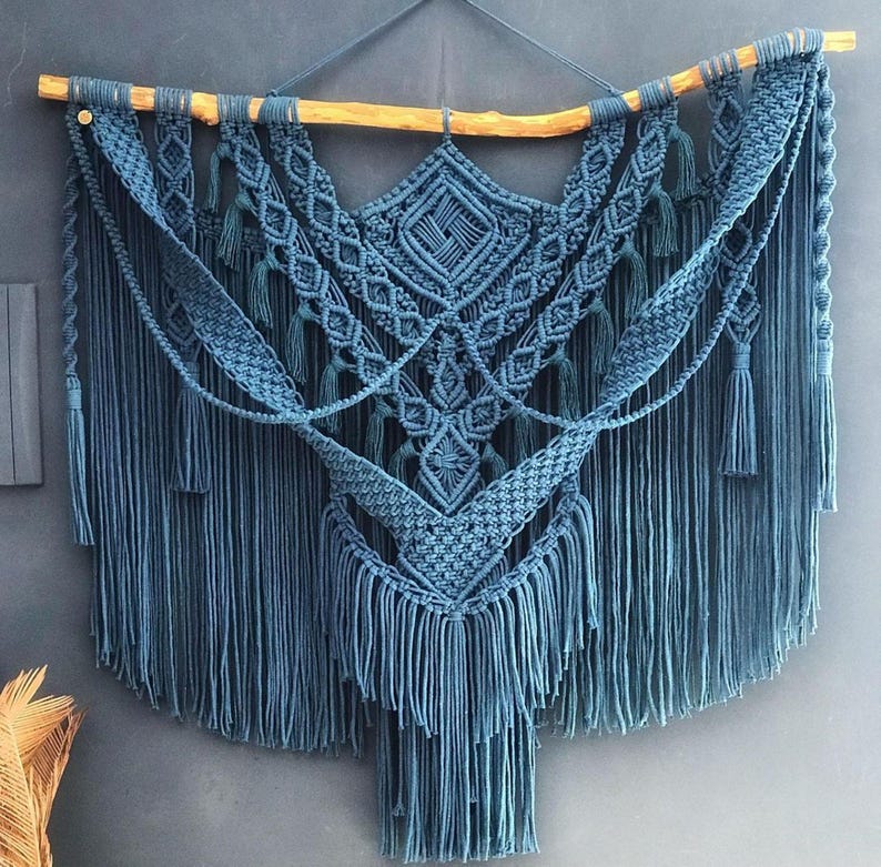 Op de afbeelding: Een grote, blauwe macram&eacute; wandhanger met een geometrisch patroon en franjes. De hanger is gemaakt van katoenen koord en is bevestigd aan een houten tak.