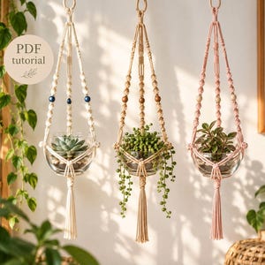 May include: Three macrame plant hangers with wooden rings at the top and tassels at the bottom. The left hanger is cream with blue beads, the center is tan with wooden beads, and the right is pink with wooden beads, each holding a glass bowl.