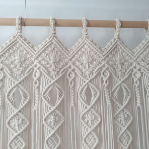 May include: Cream-colored macrame curtain with a geometric diamond pattern and fringe. The curtain is suspended from a natural wood rod. The design features intricate knotting and weaving, creating a textured, bohemian aesthetic.