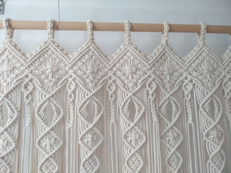 May include: Cream-colored macrame curtain with a geometric diamond pattern and fringe. The curtain is suspended from a natural wood rod. The design features intricate knotting and weaving, creating a textured, bohemian aesthetic.