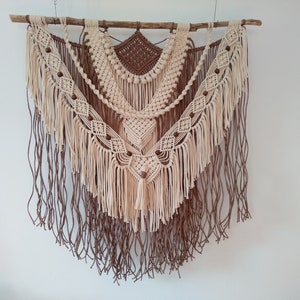 May include: A beige and brown macrame wall hanging with wooden dowel and brown wooden beads. The wall hanging features a geometric pattern with fringe.