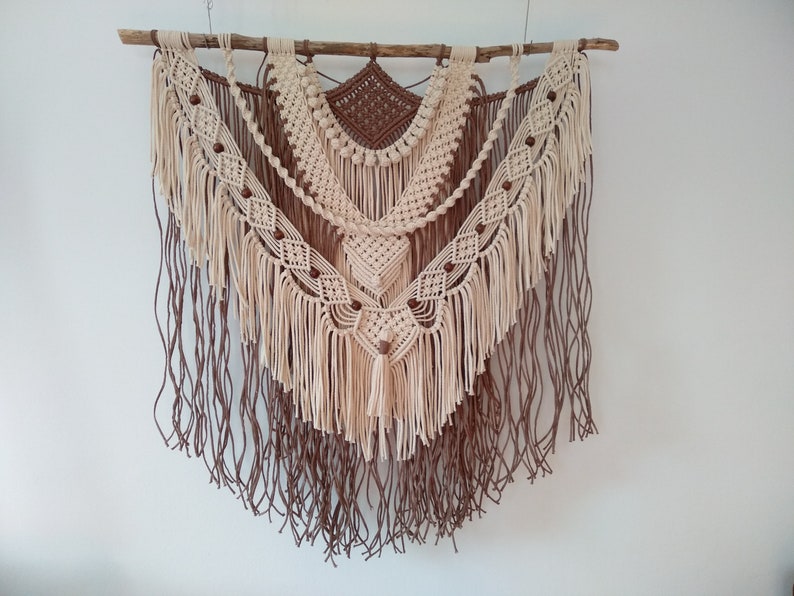May include: A beige and brown macrame wall hanging with wooden dowel and brown wooden beads. The wall hanging features a geometric pattern with fringe.