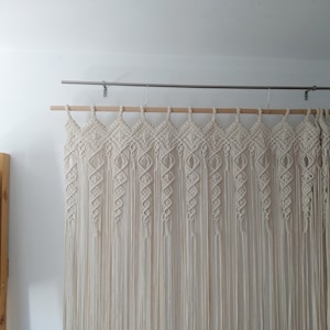 May include: Cream-colored macrame wall hanging. The textile art piece is suspended from a wooden dowel and metal rod. The design features diamond and spiral patterns, with long, vertical fringe. A wooden bookshelf with books is visible on the left.
