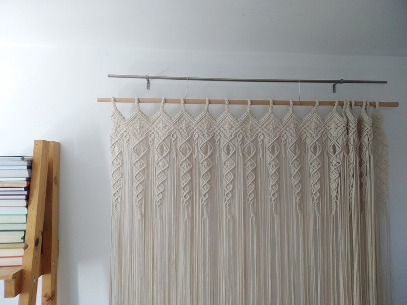 May include: Cream-colored macrame wall hanging. The textile art piece is suspended from a wooden dowel and metal rod. The design features diamond and spiral patterns, with long, vertical fringe. A wooden bookshelf with books is visible on the left.