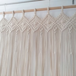 May include: A white macrame curtain with a decorative top border. The curtain is made of cotton cord and hangs from a wooden rod.
