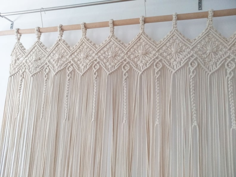 May include: A white macrame curtain with a decorative top border. The curtain is made of cotton cord and hangs from a wooden rod.