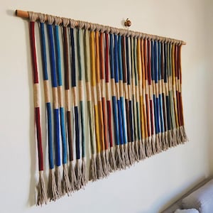 Może przedstawiać: Kolorowy wiszący dywanik ścienny wykonany z włóczki w różnych kolorach, w tym czerwonym, niebieskim, żółtym i zielonym. Włóczka jest spleciona w pasy. Dywanik jest przymocowany do drewnianej listwy.