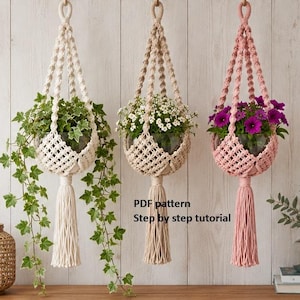 May include: Three macrame plant hangers in cream, beige, and pink, each holding a glass bowl with green ivy, white flowers, or purple flowers. Each hanger features a knotted design and a long tassel. Text reads: PDF pattern Step by step tutorial.