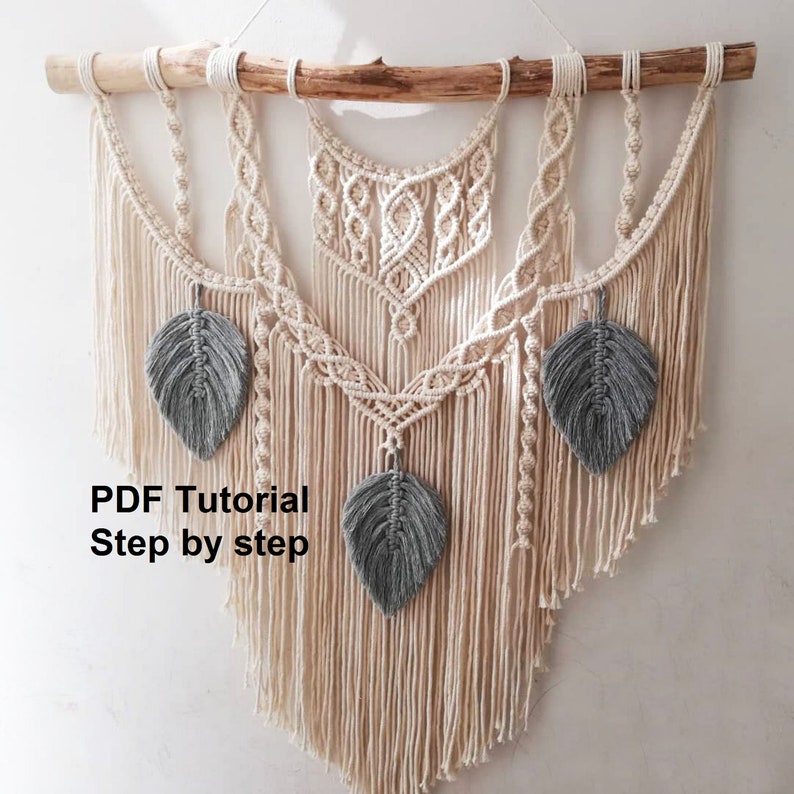 May include: A macrame wall hanging with a wooden dowel at the top and three gray feather accents. The wall hanging is made with natural colored cord and has a fringe at the bottom. The text "PDF Tutorial Step by step" is visible in the image.