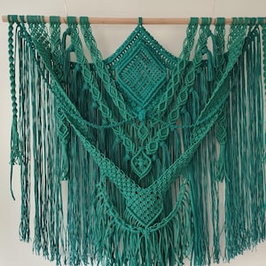 Op de afbeelding: Een tealgroene macram&eacute; wandhanger met een geometrisch ontwerp. De hanger is gemaakt van katoenen koord en heeft een franje rand.