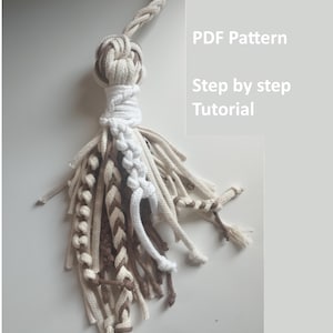Pode incluir: Uma borla de macramé com um nó central e vários cordões trançados e soltos em tons de branco, creme e castanho. O texto PDF Pattern e Step by step Tutorial é visível no lado direito, indicando um guia de instruções digital.