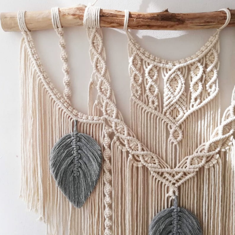 May include: A macrame wall hanging with a natural wood dowel and gray feather accents. The wall hanging features a variety of knot patterns and textures.