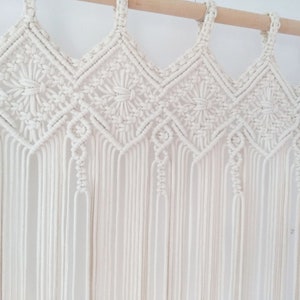 May include: A white macrame wall hanging with a geometric pattern. The hanging is made of cotton cord and has a fringe at the bottom.