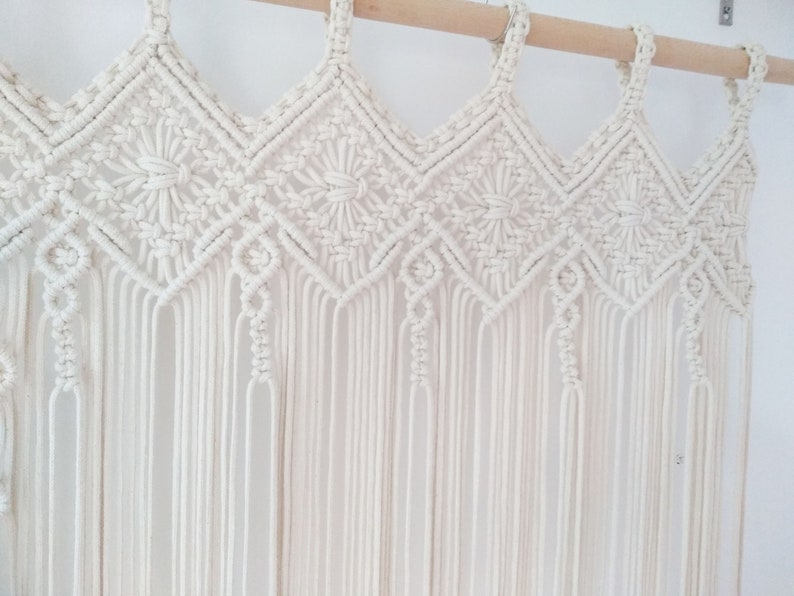 May include: A white macrame wall hanging with a geometric pattern. The hanging is made of cotton cord and has a fringe at the bottom.