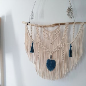 May include: A macrame wall hanging with a natural wood branch, featuring a geometric design with fringe and teal blue tassels.