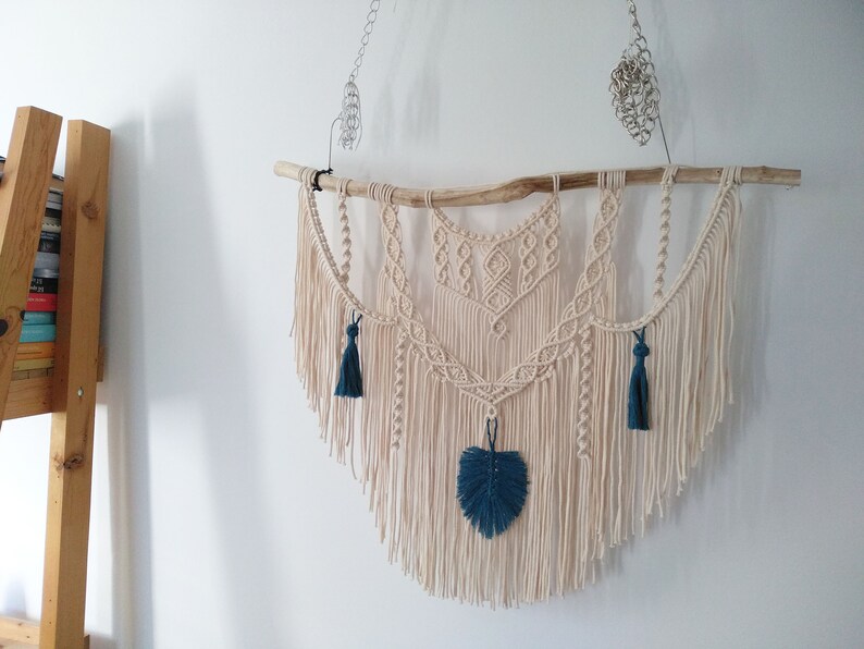 May include: A macrame wall hanging with a natural wood branch, featuring a geometric design with fringe and teal blue tassels.