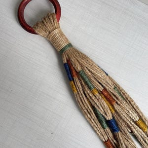 Op de afbeelding: Een bruine jute touw macrame plantenhanger met een houten ring aan de bovenkant. Het touw is versierd met blauwe, groene, oranje en bruine strepen.