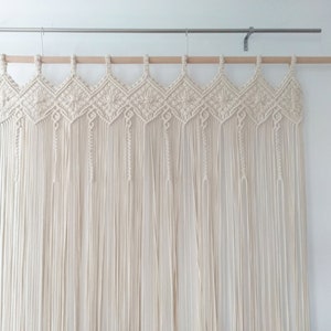 May include: A white macrame wall hanging with a geometric pattern. The hanging is made of cotton cord and is attached to a wooden dowel rod.