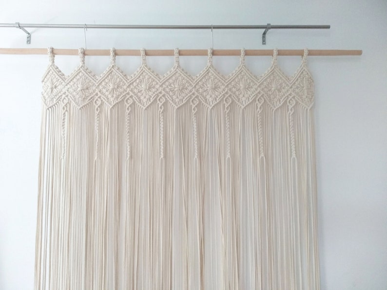 May include: A white macrame wall hanging with a geometric pattern. The hanging is made of cotton cord and is attached to a wooden dowel rod.