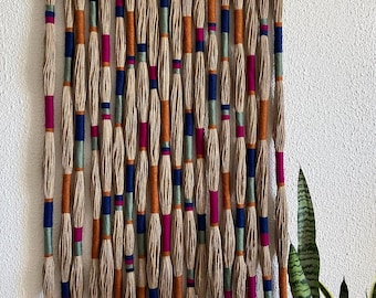 Puede incluir: Un tapiz de pared hecho de hebras de cuerda de fibra natural tejidas con acentos de hilo naranja, rosa, azul y verde.