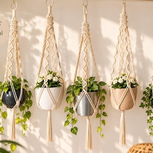 May include: Five off-white macrame plant hangers made of knotted cord, each suspending a different pot containing green foliage. The hangers feature intricate geometric patterns, circular top rings, and bottom tassels. A circular badge reads PDF tutorial.
