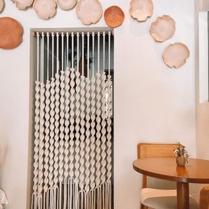 May include: A white macrame curtain hangs in a doorway, with a wooden chair and table in the foreground. The wall is decorated with several terracotta and cream-colored ceramic wall decor pieces.