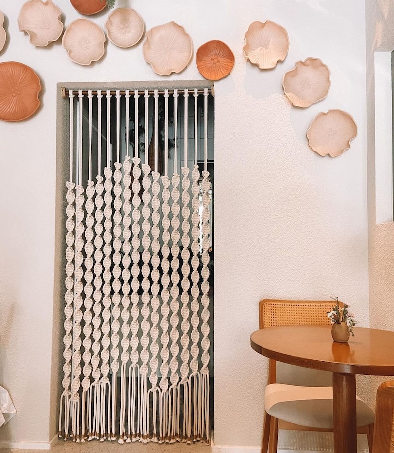 May include: A white macrame curtain hangs in a doorway, with a wooden chair and table in the foreground. The wall is decorated with several terracotta and cream-colored ceramic wall decor pieces.