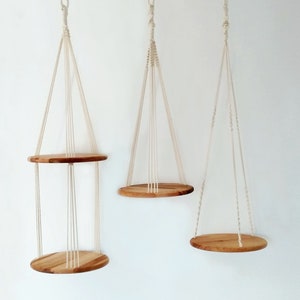 Può includere: Tre portavasi sospesi in macramè con ripiani in legno. I portavasi sono realizzati in cordoncino di cotone bianco naturale e i ripiani sono in legno marrone chiaro.