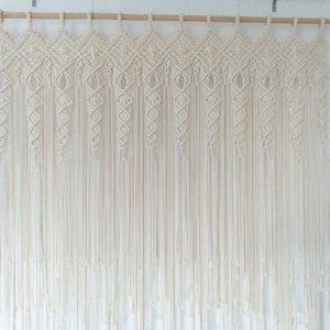 May include: Cream-colored macrame wall hanging. The top features a geometric design with diamond shapes, and the bottom has long, vertical strands. The macrame is suspended from a wooden rod, creating a bohemian aesthetic.