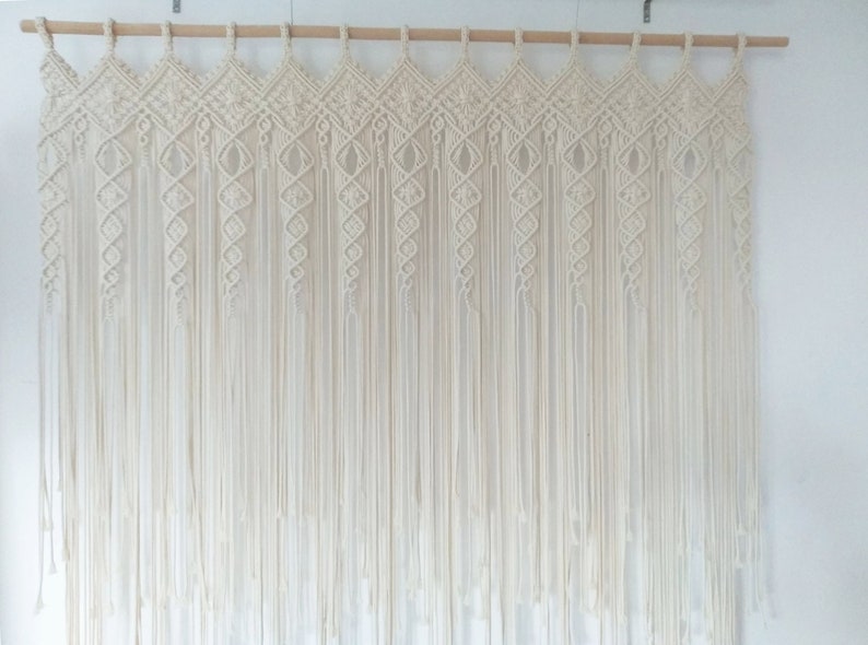 May include: Cream-colored macrame wall hanging. The top features a geometric design with diamond shapes, and the bottom has long, vertical strands. The macrame is suspended from a wooden rod, creating a bohemian aesthetic.
