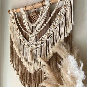 May include: A beige and brown macrame wall hanging with wooden dowel and fringe. The hanging features a geometric pattern with wooden beads.