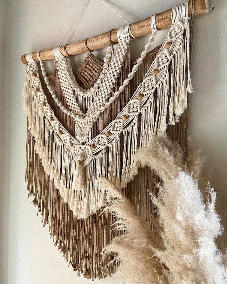 May include: A beige and brown macrame wall hanging with wooden dowel and fringe. The hanging features a geometric pattern with wooden beads.