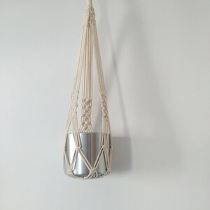 May include: A white macrame plant hanger with a silver metal pot hanging from it.