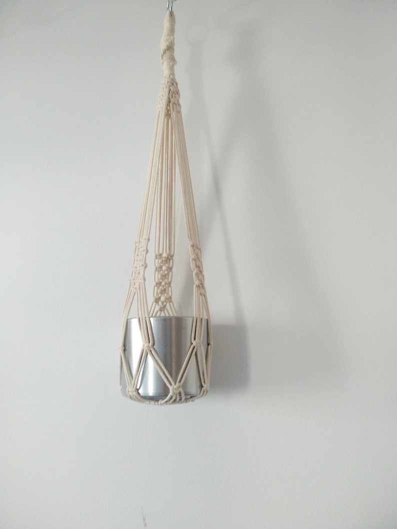 May include: A white macrame plant hanger with a silver metal pot hanging from it.