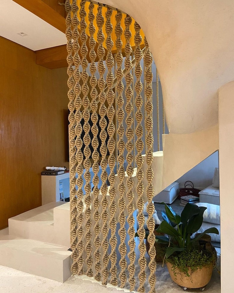 May include: A beige rope macrame wall hanging with a woven pattern. The macrame is hanging in a doorway and is partially obscured by a wooden beam.