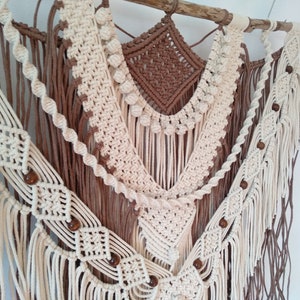 May include: A macrame wall hanging with a brown and white color scheme. The hanging features a variety of knots and patterns, including a diamond shape in the center. The hanging is decorated with wooden beads.