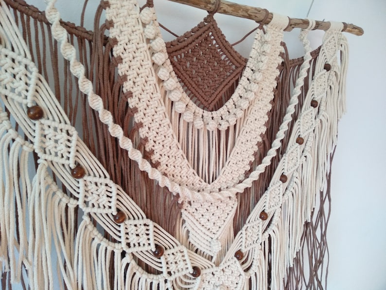 May include: A macrame wall hanging with a brown and white color scheme. The hanging features a variety of knots and patterns, including a diamond shape in the center. The hanging is decorated with wooden beads.