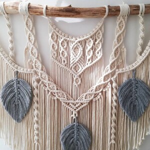 May include: A macrame wall hanging with a natural wood dowel and gray feather accents. The macrame is made with a cream-colored cord and features a variety of knots and patterns.