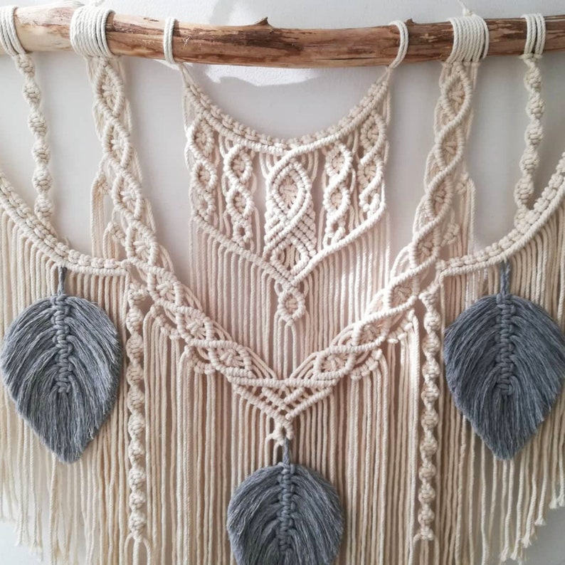 May include: A macrame wall hanging with a natural wood dowel and gray feather accents. The macrame is made with a cream-colored cord and features a variety of knots and patterns.