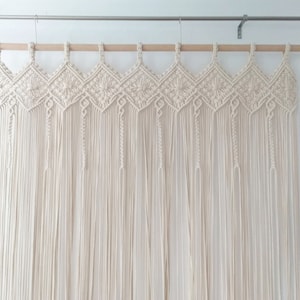 May include: A white macrame curtain with a geometric pattern hangs from a wooden rod. The curtain is made of cotton cord and has a fringe at the bottom.