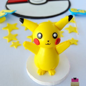 May include: A yellow Pikachu figure with red cheeks and black-tipped ears stands on a white base. A red and white Pok&eacute; Ball and yellow star decorations are in the background. The figure is a cake topper.