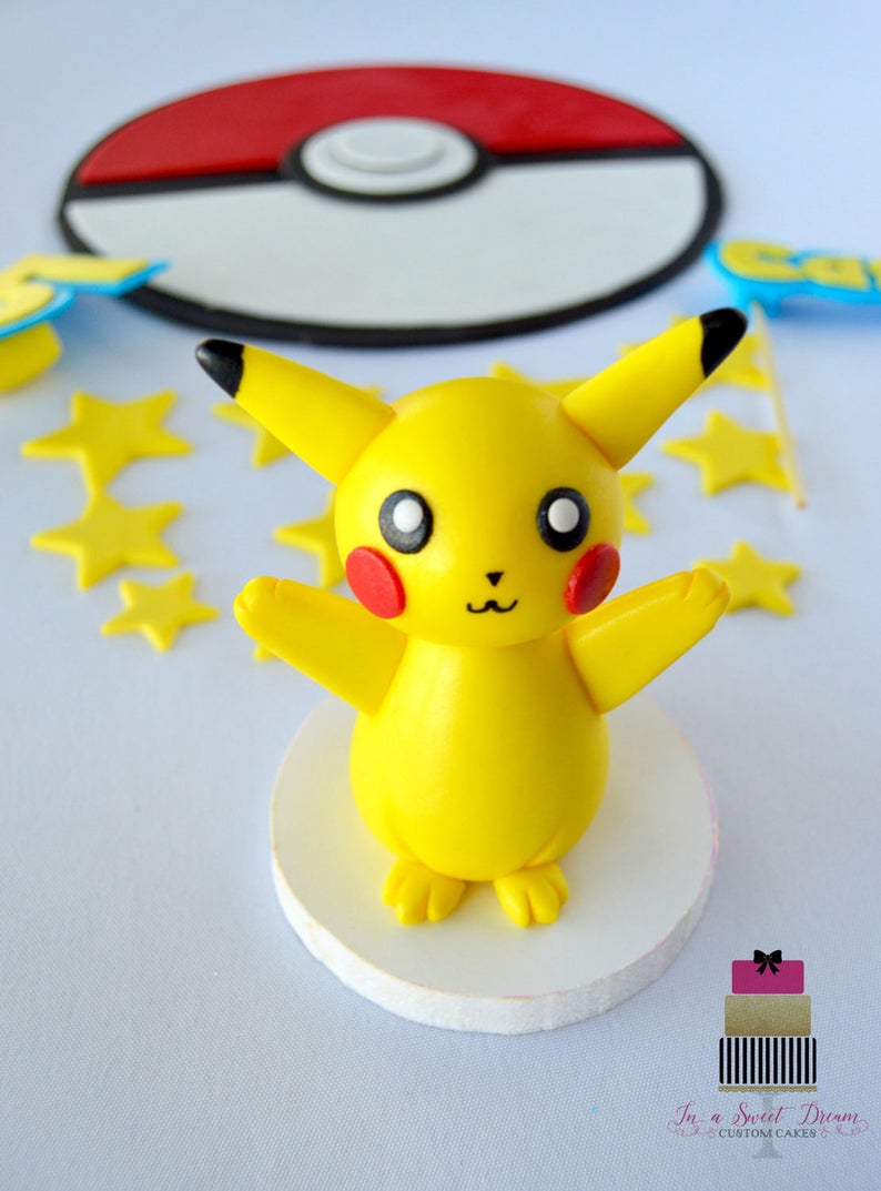 May include: A yellow Pikachu figure with red cheeks and black-tipped ears stands on a white base. A red and white Pok&eacute; Ball and yellow star decorations are in the background. The figure is a cake topper.
