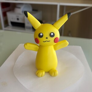 May include: A yellow Pikachu figure with black-tipped ears, red cheek circles, and a smiling mouth. The figure has outstretched arms and is standing on a white surface. The figurine is a popular character from the Pok&eacute;mon franchise.