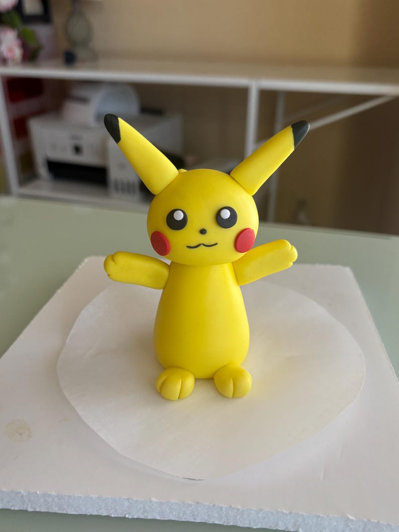 May include: A yellow Pikachu figure with black-tipped ears, red cheek circles, and a smiling mouth. The figure has outstretched arms and is standing on a white surface. The figurine is a popular character from the Pok&eacute;mon franchise.